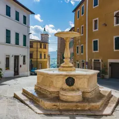 Fontana in pietra nel cuore dell'entroterra toscano, con palazzi storici e scorcio collinare.