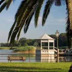 Un gazebo bianco si affaccia sul lago, vicino a una torre. Palme e panchine completano la scena del centro storico.