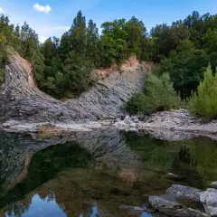 Rocce stratificate e vegetazione lussureggiante si specchiano nelle acque tranquille del Val di Farma, un paesaggio naturale mozzafiato.
