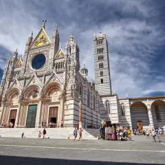 Magnifica facciata del Duomo di Siena, con la sua torre a strisce bianche e nere. Turisti passeggiano nella piazza antistante.