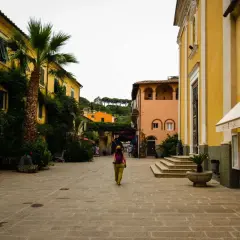 Una donna cammina in una piazza acciottolata del centro storico dell'Isola d'Elba, circondata da edifici color pastello e vegetazione lussureggiante.