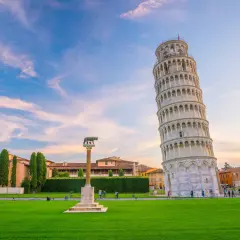 La celebre torre di Pisa, con la sua inconfondibile pendenza, si staglia contro un cielo azzurro e rosa. Un prato verde e un monumento completano la scena.