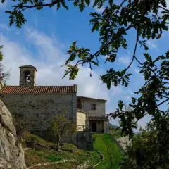 L'eremo di Cerbaiolo, con la sua pietra antica e il campanile, si staglia contro un cielo azzurro tra i rami di quercia.