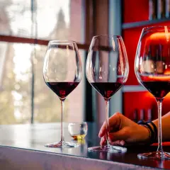 Calice di vino rosso su un bancone, con una mano che sfiora uno stelo. L'atmosfera è quella della cantina il poggio, con luci soffuse e bottiglie sullo sfondo.