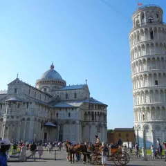 Visitare Pisa: la Torre Pendente e il Duomo svettano sotto un cielo azzurro, mentre turisti e una carrozza trainata da cavalli animano la piazza.