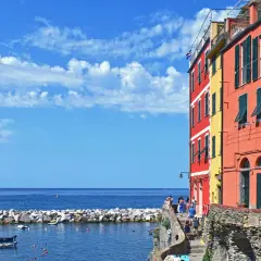 Case, a Firenze c'è il mare! Case colorate affacciate su un porto con barche, sotto un cielo azzurro con nuvole bianche.