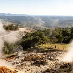 Vapori emergono dal terreno in un paesaggio collinare, con un villaggio in lontananza. Un geyser in Italia che incanta i visitatori.