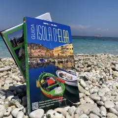 Guida turistica "Isola d'Elba" su una spiaggia di ciottoli, con il mare azzurro sullo sfondo. Perfetta per pianificare la tua vacanza.