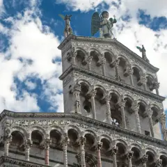 La facciata riccamente decorata della chiesa San Michele a Lucca, con un angelo in cima, si staglia contro un cielo azzurro e nuvoloso.