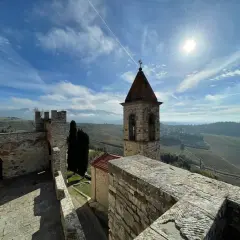 Torre campanaria e mura antiche del castello Frescobaldi, con vigneti e colline toscane sotto un cielo azzurro e soleggiato.