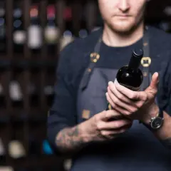 Un sommelier mostra una bottiglia di **vini toscani rossi pregiati**, con scaffali pieni di bottiglie sullo sfondo.