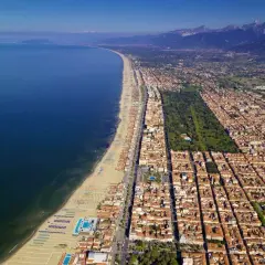 Vista aerea della costa di Massa Carrara, con il mare blu intenso, la lunga spiaggia attrezzata e la città che si estende verso le montagne.