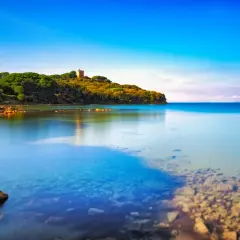 La costa verdeggiante con una torre antica, dove si trova Punta Ala, si specchia nelle acque calme e cristalline del mare.