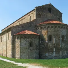 Basilica romanica di San Piero a Grado, con absidi semicircolari e tetto in tegole rosse, su un prato verde sotto un cielo azzurro.