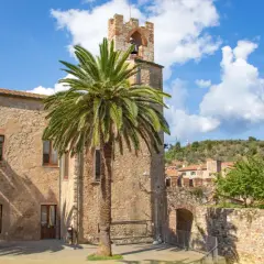 Un antico campanile in pietra a Suvereto, Livorno, svetta tra palme rigogliose e un cielo azzurro con nuvole bianche.