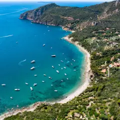 Baia con acque cristalline e numerose barche, incastonata tra la costa verdeggiante e le pendici del monte Argenia.
