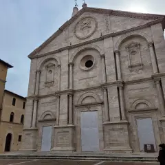 Facciata imponente del Duomo di Pienza, con accanto un edificio in pietra che ricorda il Palazzo Piccolomini.