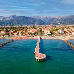 Un molo si estende nel mare turchese di Marina di Massa, con spiagge affollate e montagne sullo sfondo.