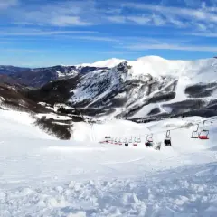 Impianti di risalita rossi trasportano sciatori su piste innevate ad Abetone, con montagne sullo sfondo sotto un cielo azzurro.