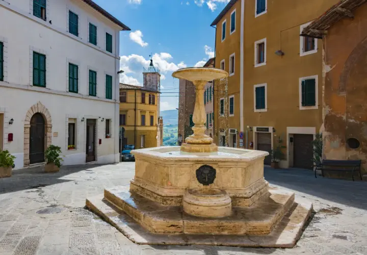 Fontana in pietra nel cuore dell'entroterra toscano, con palazzi storici e scorcio collinare.