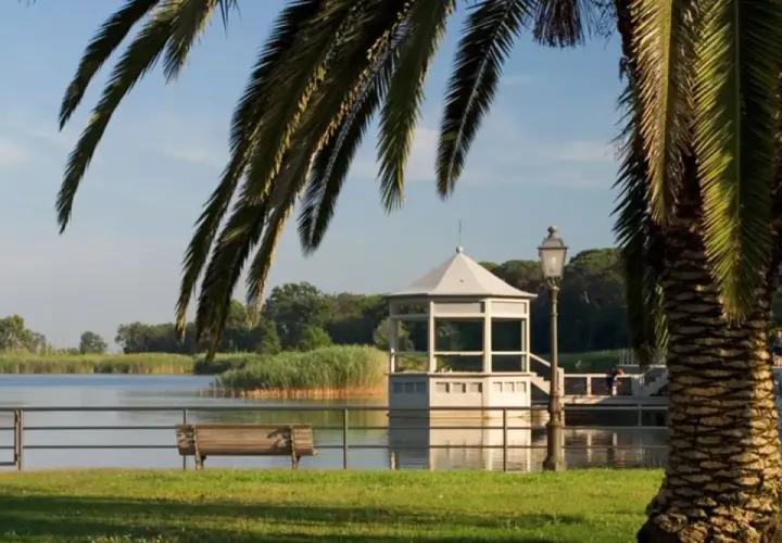 Un gazebo bianco si affaccia sul lago, vicino a una torre. Palme e panchine completano la scena del centro storico.