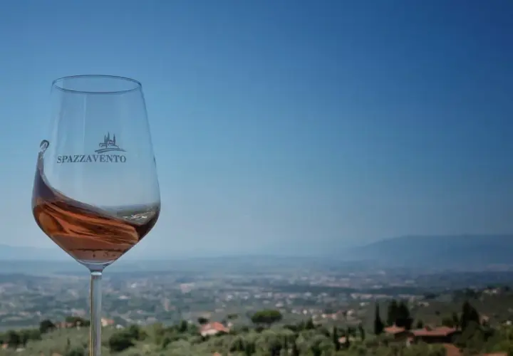 Calice di vino rosato con logo Spazzavento, sullo sfondo il panorama della fattoria di Piazzano e le colline toscane.