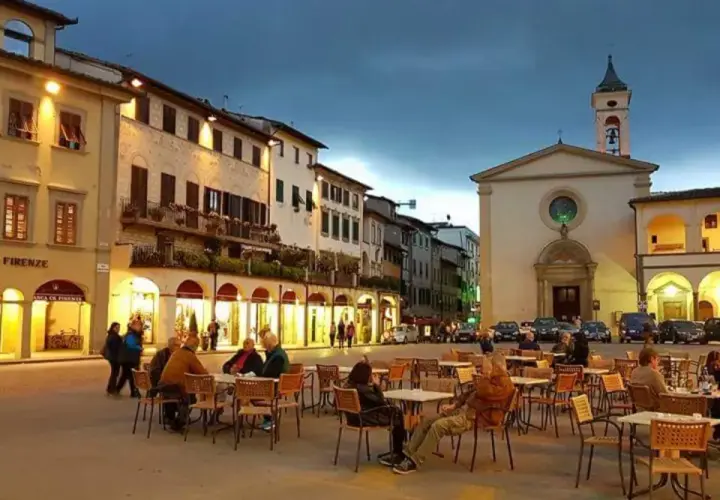 Serata in piazza a Figline Valdarno, con tavolini all'aperto e la chiesa illuminata.