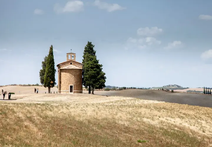 Iconica chiesetta Val d'Orcia, con cipressi e colline ondulate sotto un cielo nuvoloso.