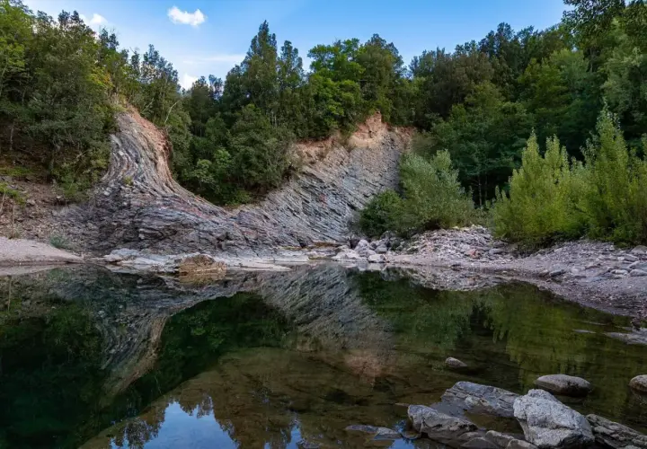 Rocce stratificate e vegetazione lussureggiante si specchiano nelle acque tranquille del Val di Farma, un paesaggio naturale mozzafiato.