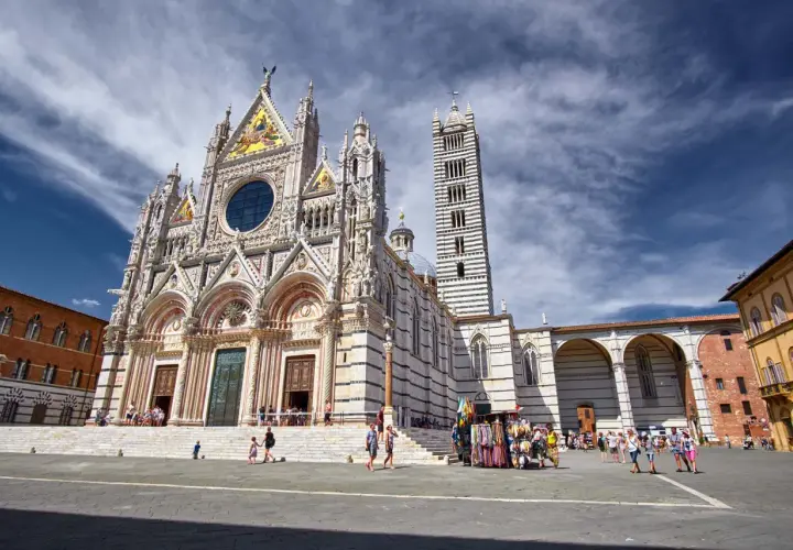 Magnifica facciata del Duomo di Siena, con la sua torre a strisce bianche e nere. Turisti passeggiano nella piazza antistante.