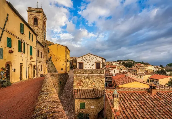 Un vicolo con case gialle e torre campanaria a Montescudaio. Cosa vedere: un borgo medievale con tetti rossi e cielo nuvoloso.