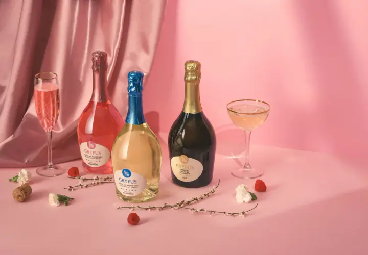 Tre bottiglie di champagne italiano, una rosata, una bianca e una dorata, con calici e decorazioni floreali su sfondo rosa.