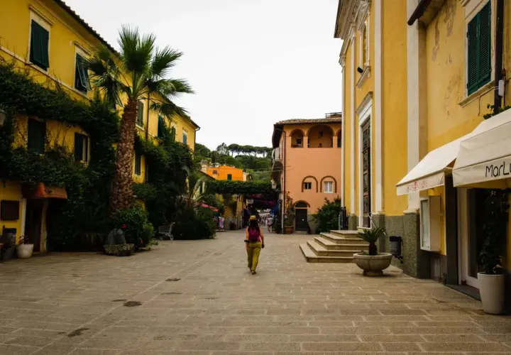 Una donna cammina in una piazza acciottolata del centro storico dell'Isola d'Elba, circondata da edifici color pastello e vegetazione lussureggiante.