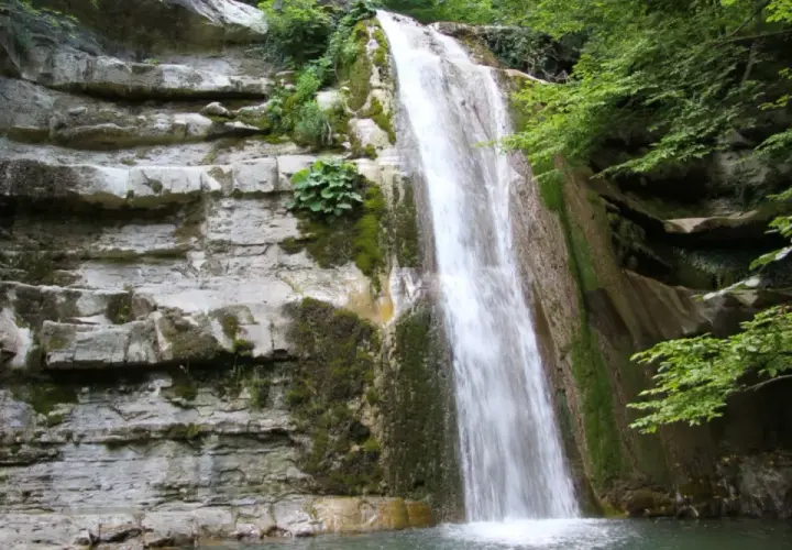 Cascata fragorosa su rocce stratificate, circondata da vegetazione lussureggiante. L'acqua cheta scende impetuosa, creando un velo bianco.
