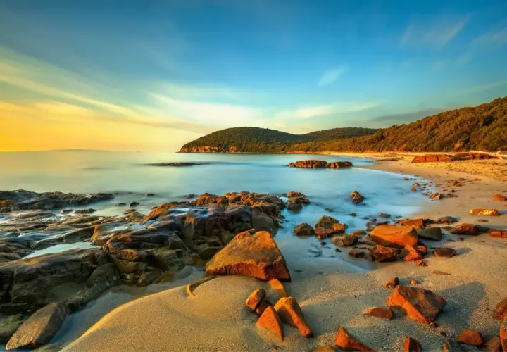 La Cala Martina al tramonto, con rocce scure e sabbia dorata bagnata da un mare calmo e cielo sfumato.