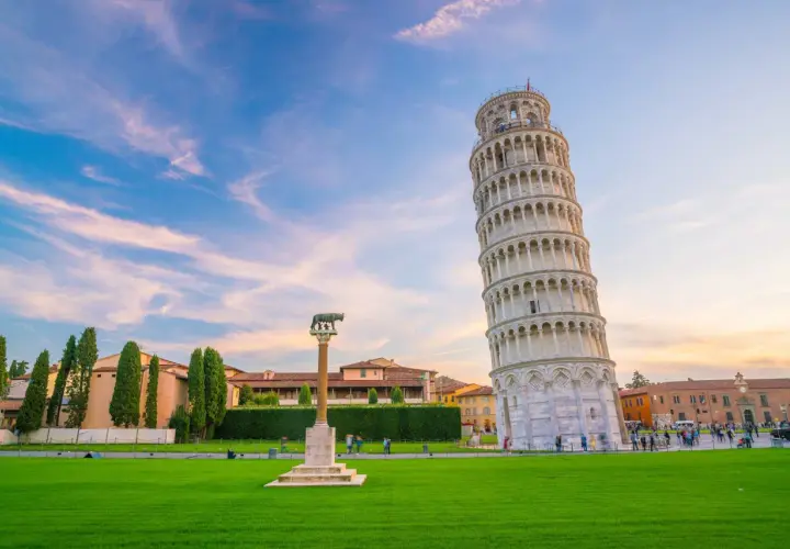 La celebre torre di Pisa, con la sua inconfondibile pendenza, si staglia contro un cielo azzurro e rosa. Un prato verde e un monumento completano la scena.