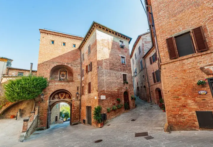 Antica porta in mattoni a Torrita di Siena, con un affresco e un vicolo acciottolato che sale tra case medievali.