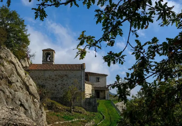 L'eremo di Cerbaiolo, con la sua pietra antica e il campanile, si staglia contro un cielo azzurro tra i rami di quercia.