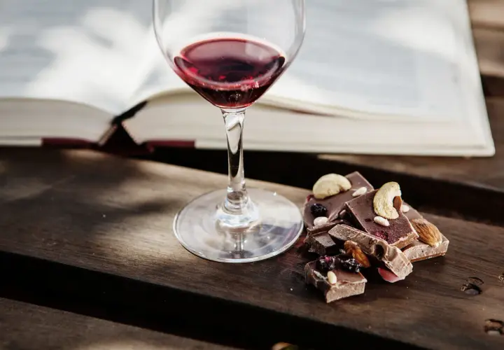 Un bicchiere di vino rosso accanto a pezzi di cioccolato con frutta secca e noci. Un perfetto abbinamento vino e cioccolato per una serata di relax.