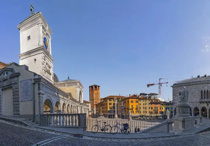 La piazza della Libertà, con la sua torre dell'orologio, edifici colorati e due biciclette in primo piano, sotto un cielo azzurro.