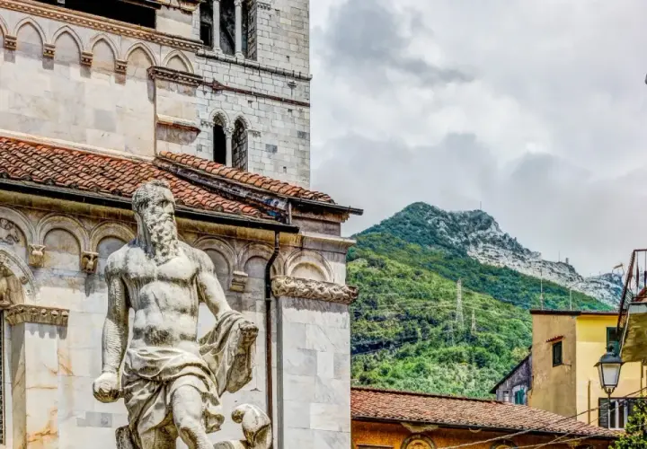 Statua nel carrara centro storico, con montagne verdi sullo sfondo e case colorate.