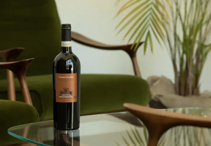 Bottiglia di vino rosso Poggio Miniera, un'eccellenza tra i vini toscani, su un tavolino di vetro, con poltrone verdi e piante sullo sfondo.