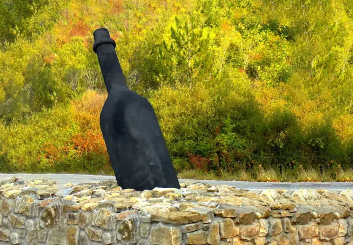 Giant black wine bottle sculpture emerging from a stone wall, with a colorful autumn forest in the background.
