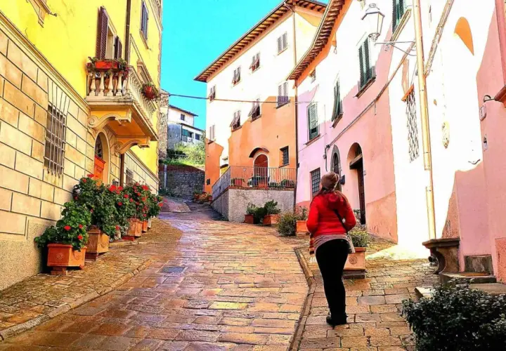 Una donna cammina su una strada acciottolata a Palaia, Pisa, tra case colorate e vasi di fiori rossi.