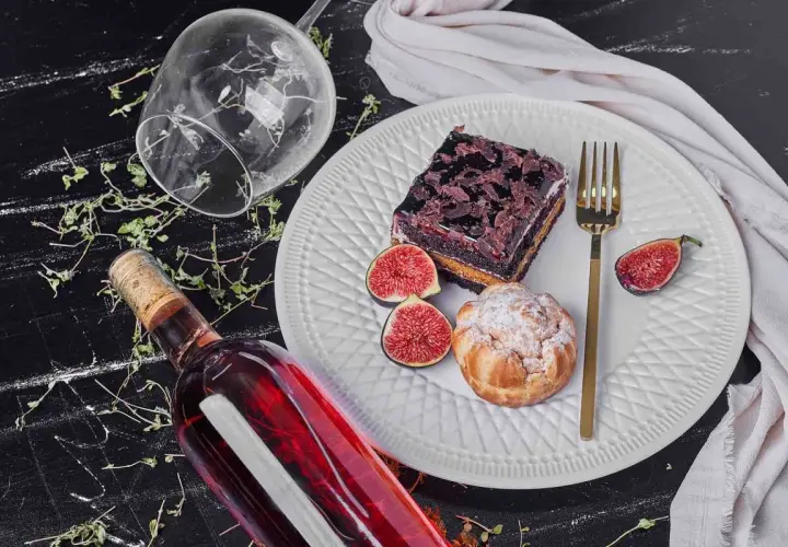 Un bicchiere rovesciato, una bottiglia di vino rosso e un piatto con torta al cioccolato, bignè, fichi e posate dorate, un'invitante composizione per gustare vini da dessert.