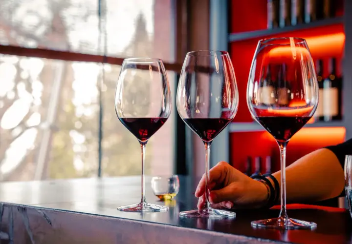 Calice di vino rosso su un bancone, con una mano che sfiora uno stelo. L'atmosfera è quella della cantina il poggio, con luci soffuse e bottiglie sullo sfondo.