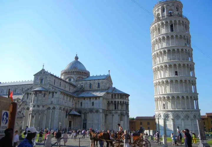Visitare Pisa: la Torre Pendente e il Duomo svettano sotto un cielo azzurro, mentre turisti e una carrozza trainata da cavalli animano la piazza.