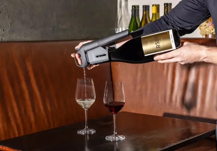 Versando vino bianco da una bottiglia con un erogatore Coravin, accanto a un bicchiere di vino rosso. Un tipo di vino da scoprire.