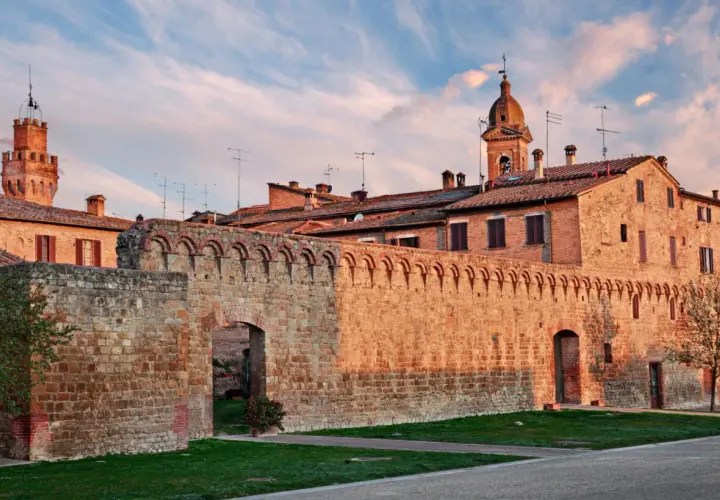 Antica cinta muraria in pietra delle Crete Senesi, con torri medievali e cielo al tramonto.