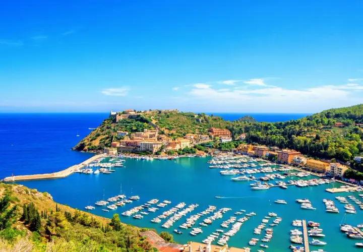Porto Ercole cosa vedere: un incantevole porto con barche, case colorate e un castello su una collina verde, bagnato da un mare azzurro.